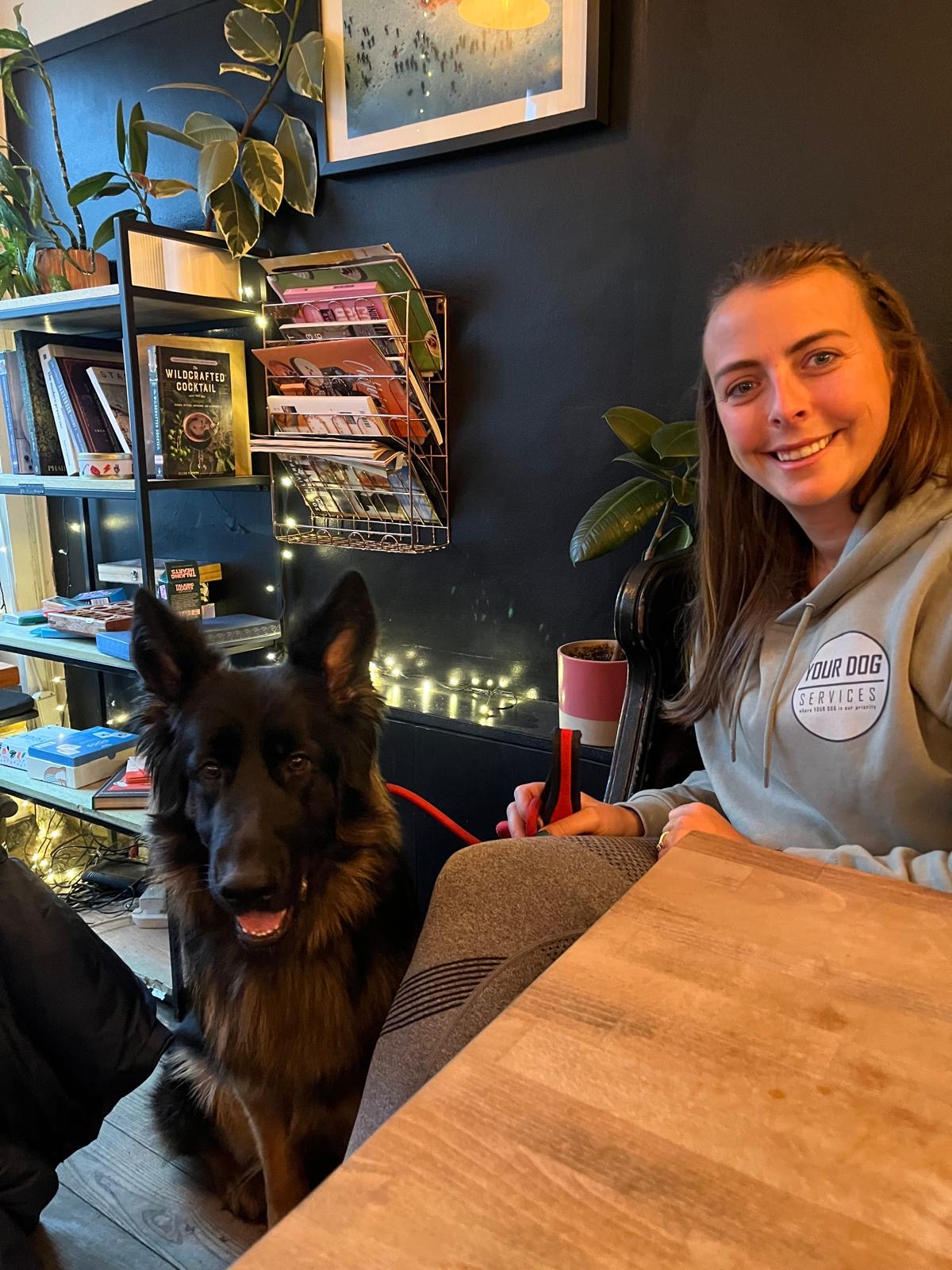 Laura, founder of Your Dog Services, in her team hoodie with a German Shepherd at a café